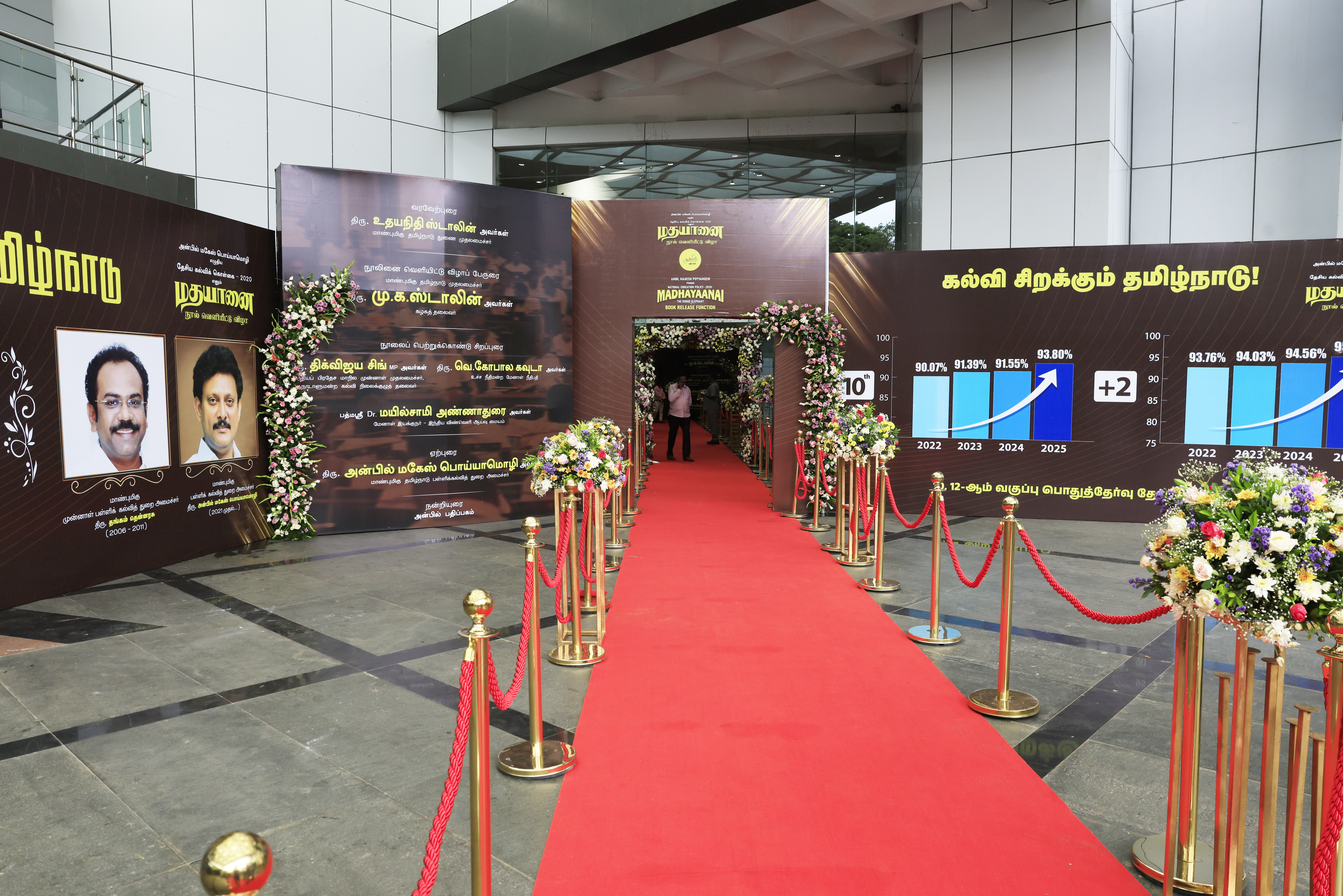 Red carpet entrance to the book release of "NEP 2020: Madhayaanai", flanked by floral arrangements, charts, and panels commemorating Tamil Nadu’s educational progress.